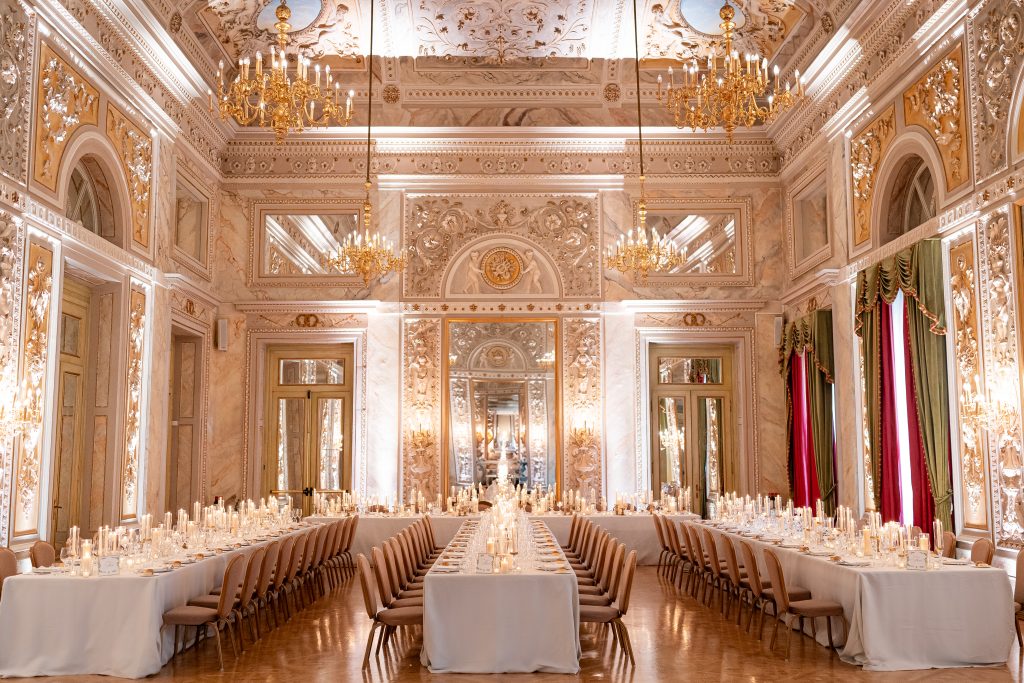 In questa foto si vede una delle tendenze allestimenti location matrimonio 2026: un salone storico decorato in stile barocco, con pareti intarsiate, specchi dorati, soffitti affrescati e lampadari a braccio. I tavoli imperiali sono disposti a ferro di cavallo, illuminati da centinaia di candele che creano un’atmosfera calda e romantica.