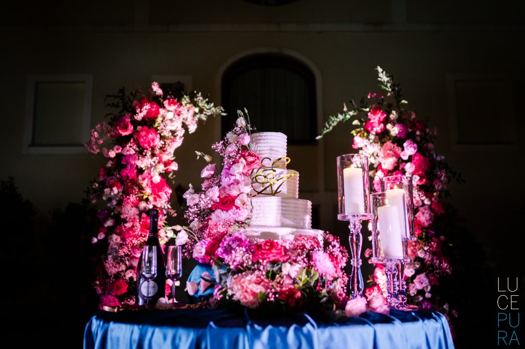 In questa foto si vede una delle tendenze allestimenti location matrimonio 2026: un tavolo per il taglio torta all’aperto, con base in tessuto blu elettrico, decorato con fiori fucsia e rosa in abbondanza. La torta è bianca e a più piani, incorniciata da archi floreali intensi e da alte candele cilindriche in vetro, creando un forte impatto visivo notturno.