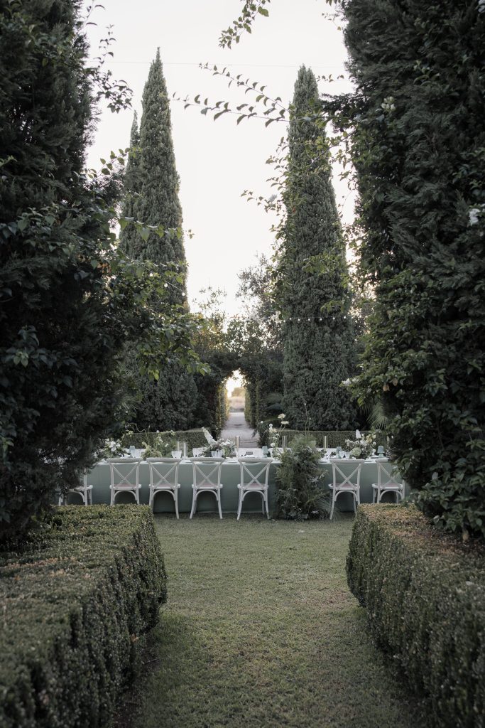 In questa foto si vede una delle tendenze allestimenti location matrimonio 2026: un lungo tavolo immerso in un giardino all’italiana, circondato da siepi geometriche e alberi ad alto fusto. Le sedie bianche in stile country chic e il verde dominante del paesaggio trasmettono un senso di intimità e armonia naturale.