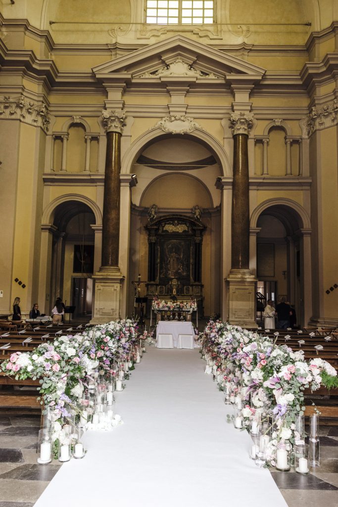 In questa foto si vede una delle tendenze allestimenti location matrimonio 2026: una navata di chiesa classica allestita con un tappeto bianco e due file di composizioni floreali abbondanti nei toni rosa, lilla e bianco. Le composizioni sono accompagnate da candele cilindriche in vetro, creando un percorso luminoso e profumato verso l’altare.