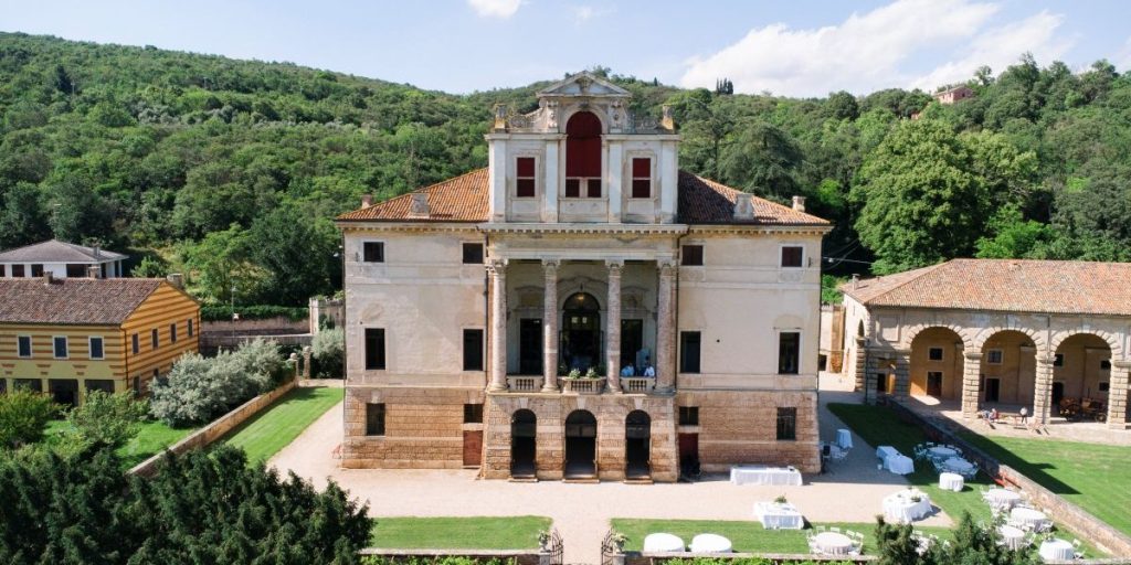 In questa foto si vede uno dei momenti di un matrimonio intimo Villa Francanzan Piovene: la storica villa veneta immersa nel verde dei Colli Berici si staglia maestosa al centro dell’immagine, con la sua facciata settecentesca in pietra e colonne corinzie. In primo piano, il giardino allestito con tavoli rotondi bianchi e sedute disposte per accogliere un ricevimento elegante e raccolto. L’atmosfera è quella di una celebrazione raffinata, immersa nella natura e nella bellezza senza tempo di un luogo autentico.