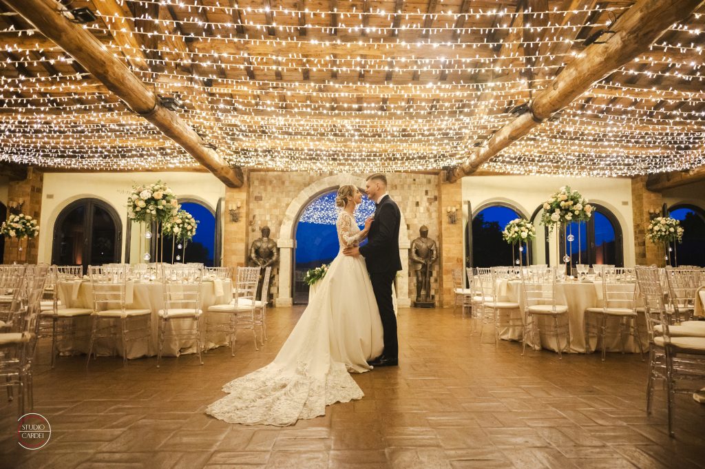 In questa foto si vede una coppia di sposi al centro di una sala ricevimenti con soffitto in legno decorato da centinaia di lucine calde sospese, mentre si abbracciano circondati da tavoli elegantemente apparecchiati e composizioni floreali alte, in un matrimonio curato da Chiara di Bari.