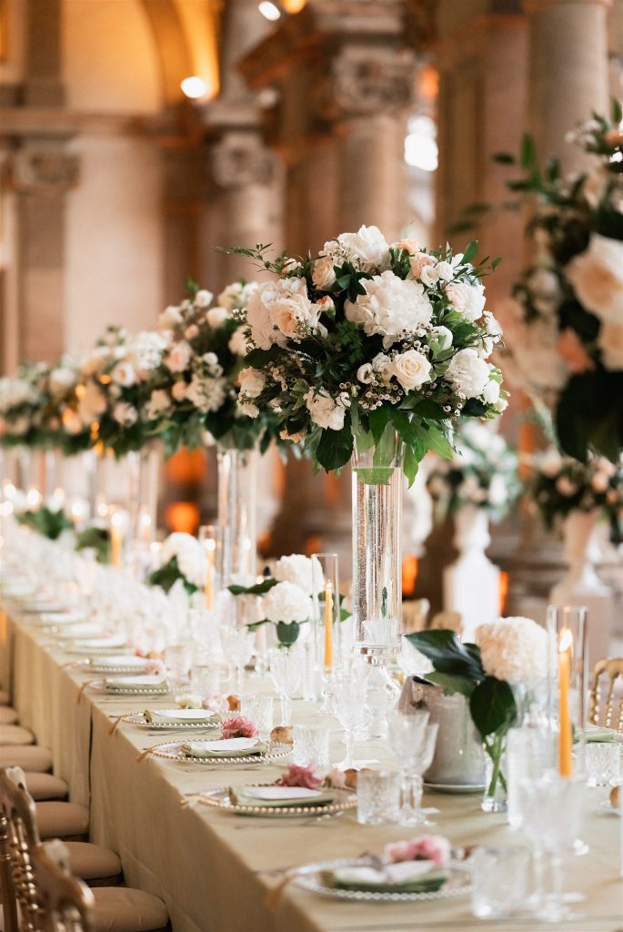 In questa foto si vede un lungo tavolo imperiale decorato con centrotavola alti in vetro trasparente, ricchi di rose bianche e verde fresco, accompagnati da candele sottili accese che creano un’atmosfera calda e raffinata in una location storica, allestimento firmato Chiara di Bari.