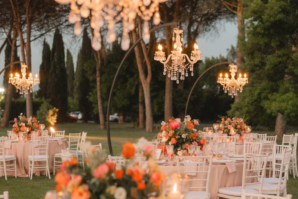 In questa foto si vede una cena all’aperto al tramonto con tavoli rotondi decorati da composizioni floreali nei toni arancio e corallo, illuminati da eleganti lampadari sospesi su strutture circolari nere, in un ricevimento serale progettato da Chiara di Bari.