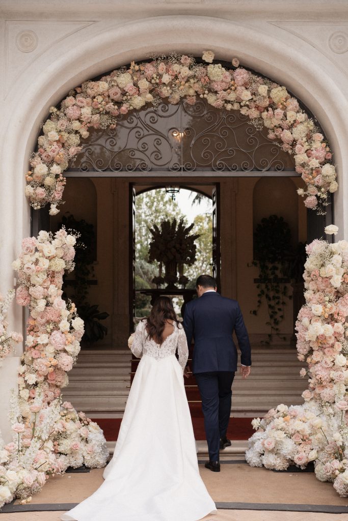In questa foto si vede una grande arcata d’ingresso completamente incorniciata da un’installazione floreale rosa cipria e bianca, mentre gli sposi entrano mano nella mano nella location, esempio della cura scenografica tipica di Chiara di Bari.