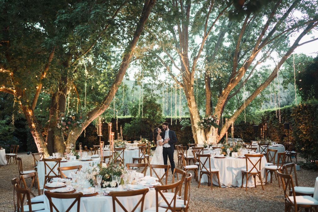 In questa foto si vede una coppia di sposi al centro di un giardino illuminato da fili di luci sospese tra gli alberi, circondati da tavoli rotondi con sedie in legno e composizioni naturali, in un’atmosfera romantica ideata da Chiara di Bari.