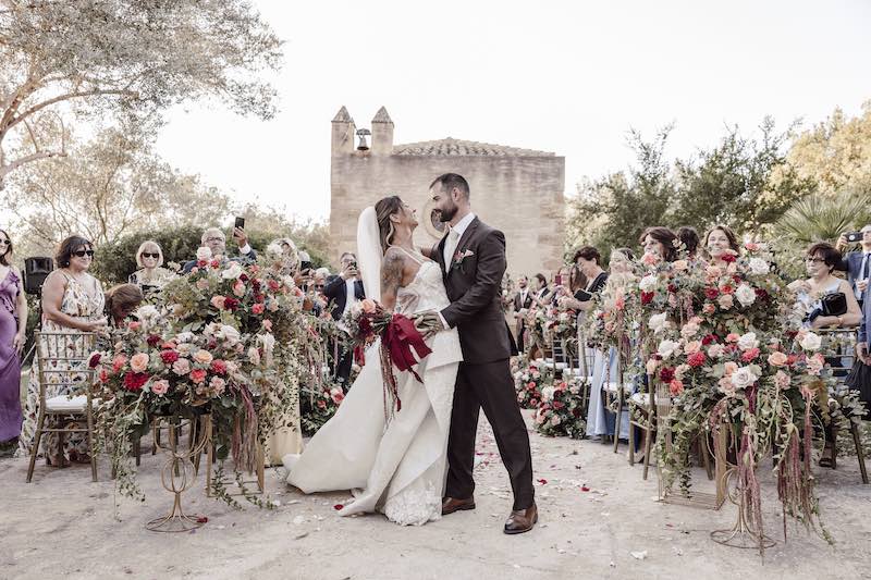 In questa foto, due sposi abbracciati alla fine della cerimonia di nozze, circondati da composizioni floreali barocche mentre dietro si intraveder la facciata di una chiesetta