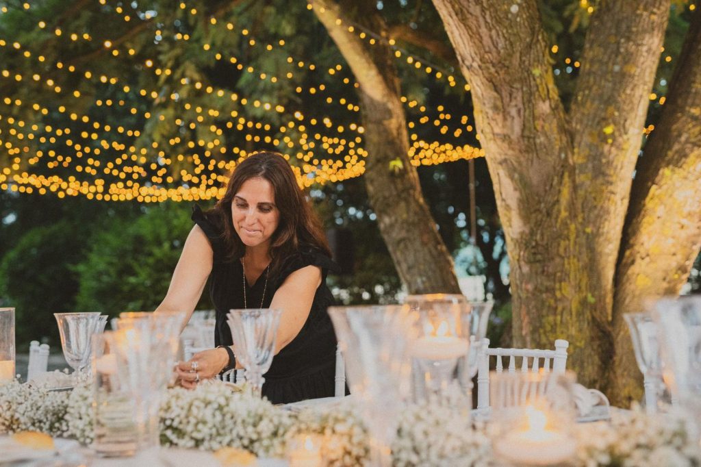 In questa foto si vede Chiara di Bari concentrata nell’allestimento di una mise en place sotto alberi decorati con luci calde sospese, mentre dispone con precisione candele e composizioni floreali bianche su una tavola raffinata in un giardino serale.