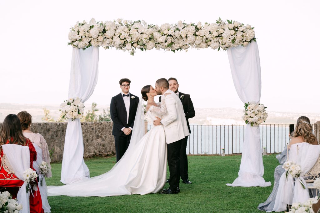 In questa foto si vede gli sposi durante il rito civile all’aperto mentre si scambiano un bacio sotto un arco floreale bianco decorato con ortensie e tessuti leggeri, con lago e panorama sullo sfondo, in una cerimonia organizzata da Chiara di Bari.