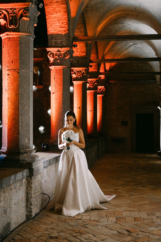 In questa foto si vede la sposa che cammina in un porticato con colonne illuminate da luci calde arancioni, indossando un abito bianco con corpetto strutturato e bouquet candido, in un contesto scenografico coordinato da Chiara di Bari.