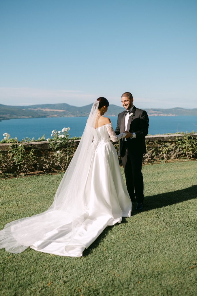 In questa foto si vede gli sposi durante la cerimonia all’aperto con vista panoramica su un lago, mentre si tengono per mano su un prato verde, circondati da composizioni floreali bianche e sedie in ferro battuto, in un evento progettato da Chiara di Bari.