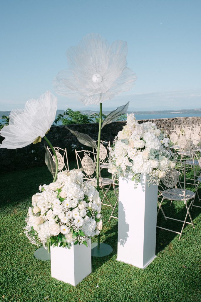 In questa foto si vede un allestimento cerimonia all’aperto con grandi installazioni floreali bianche su piedistalli, arricchite da elementi scenografici a forma di fiore oversize, affacciate su un panorama con lago sullo sfondo, esempio dello stile progettuale firmato Chiara di Bari.