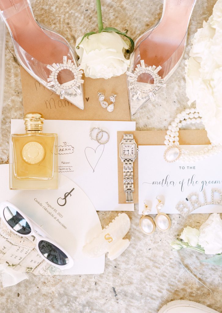 In questa foto si vede un flat lay degli accessori della sposa, con scarpe trasparenti gioiello, profumo, gioielli, orologio, occhiali da sole bianchi e partecipazioni coordinate, disposti su una superficie chiara, esempio della cura stilistica e grafica tipica di Chiara di Bari.