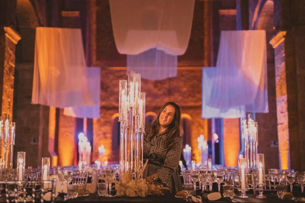 In questa foto si vede Chiara di Bari all’interno di una location storica illuminata da luci scenografiche arancioni e blu, mentre controlla un’imponente composizione di candelabri in vetro al centro di una sala con arcate in pietra e tavoli elegantemente apparecchiati.