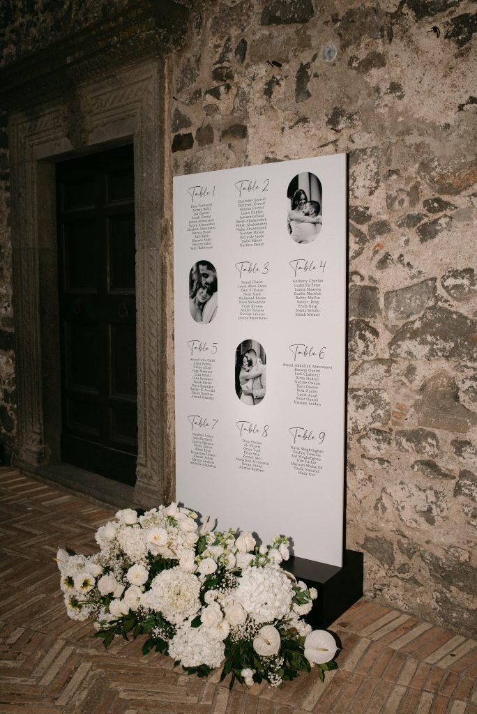 In questa foto si vede un tableau de mariage minimalista con l’elenco dei tavoli e le fotografie degli sposi, appoggiato su una parete in pietra antica e decorato alla base con una ricca composizione di fiori bianchi, in linea con l’eleganza coordinata di Chiara di Bari.