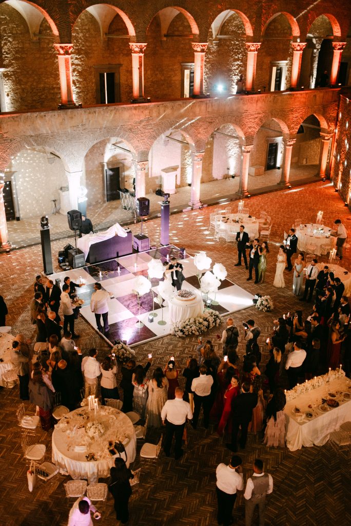 In questa foto si vede il momento del taglio torta durante il ricevimento serale in un chiostro illuminato da luci calde e architetture storiche, con gli invitati disposti intorno alla pista da ballo bianca e nera e decorazioni floreali bianche firmate Chiara di Bari.