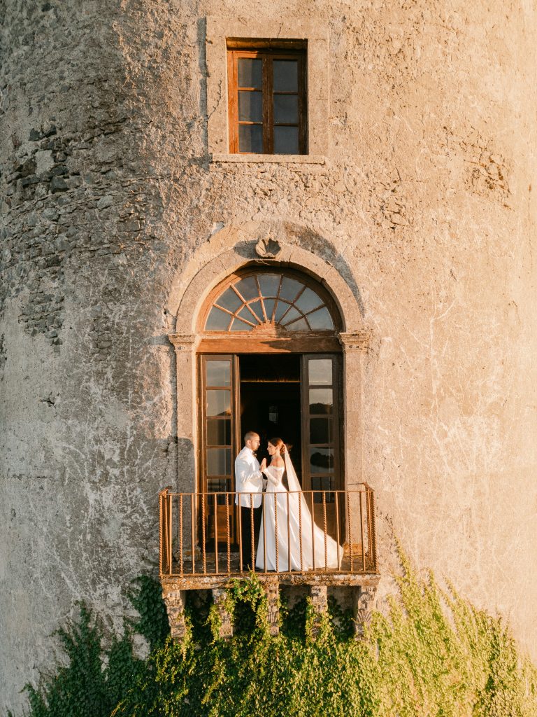 In questa foto si vede una coppia di sposi affacciata a un balcone in pietra di una dimora storica, con la sposa in abito bianco e velo lungo e lo sposo in abito chiaro, immersi nella luce dorata del tramonto, in un matrimonio organizzato da Chiara di Bari.