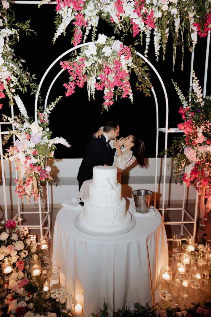 In questa foto, due sposi si baciano al taglio della torta nuziale bianca circondati da fiori di colore rosa e bianco