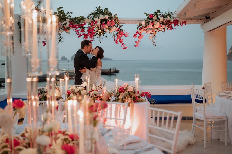In questa foto, due sposi si baciano nel luogo del loro ricevimento di nozze a Panarea, alla luce del tramonto, tra candele e fiori di colore bianco e fucsia