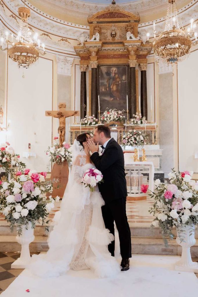 In questa foto, due sposi si baciano al termine della cerimonia di nozze nella chiesa di San Pietro a Panarea, circondati da fiori nei toni del bianco e del fucsia