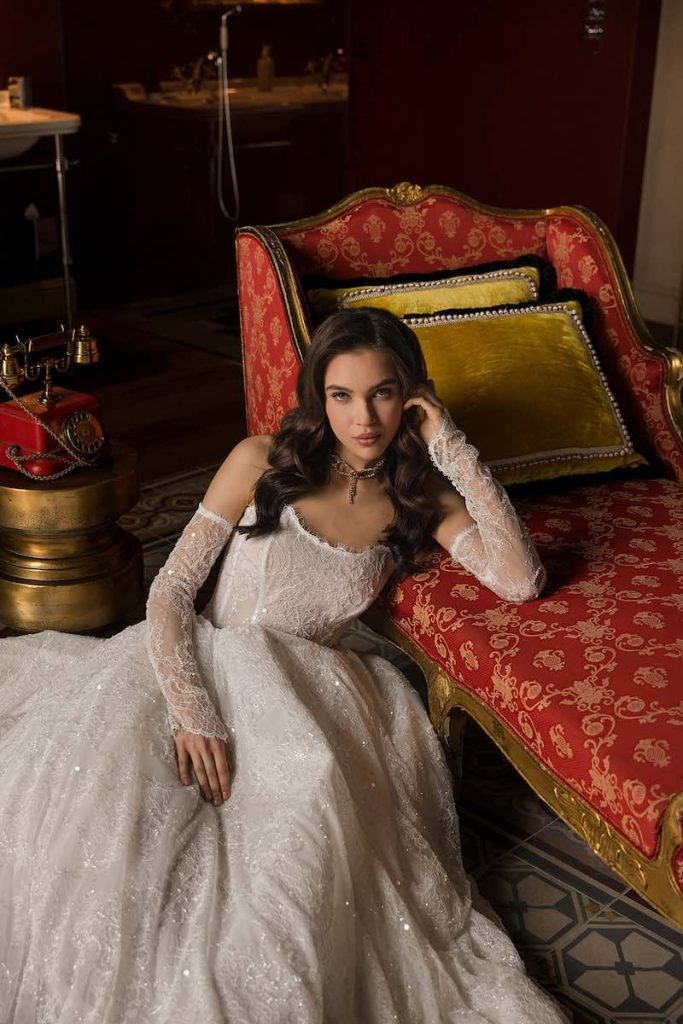 In questa foto una modella posa seduta indossando un abito da sposa Donatella Gallo 2027, ampio, con ricami preziosi e manicotti abbinati
