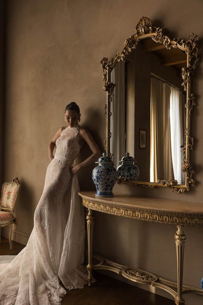 In questa foto, una modella posa indossando un abito da sposa a sirena in pizzo Donatella Gallo 2027 con scollo all'americana e sopragonna con lungo strascico in pizzo