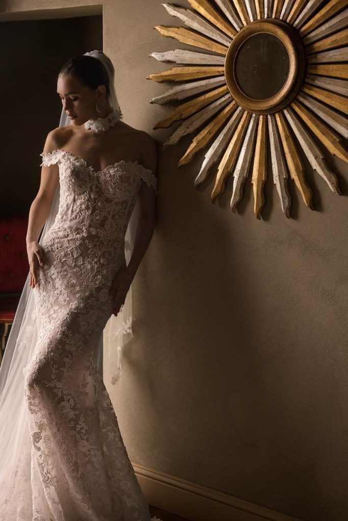 In questa foto, il dettaglio una modella che posa indossando un abito da sposa a sirena in pizzo Donatella Gallo 2027 con scollo omerale in pizzo con lungo velo abbinato