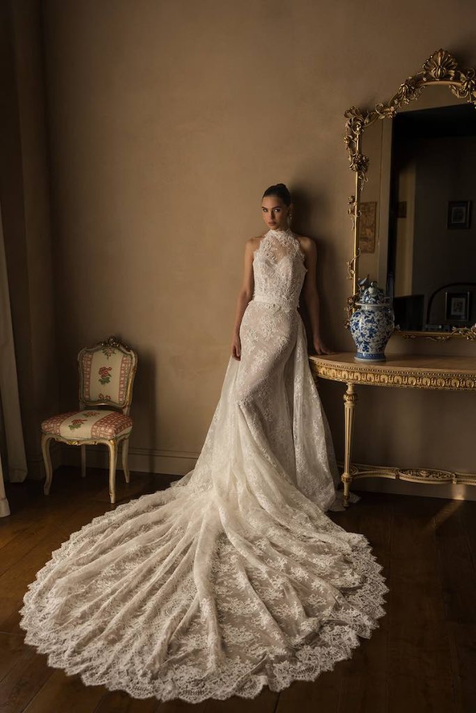 In questa foto, una modella posa indossando un abito da sposa a sirena in pizzo Donatella Gallo 2027 con scollo all'americana e sopragonna con lungo strascico in pizzo