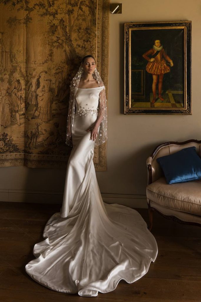 In questa foto, una modella posa indossando un abito da sposa in seta con lungo strascico, con ricami preziosi in vita e un velo con applicazioni 3D