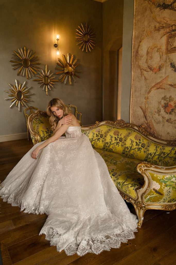 In questa foto, una modella posa seduta su un divano antico in un casale indossando un abito da sposa ad A con gonna in pizzo ed organza e un corpetto strutturato