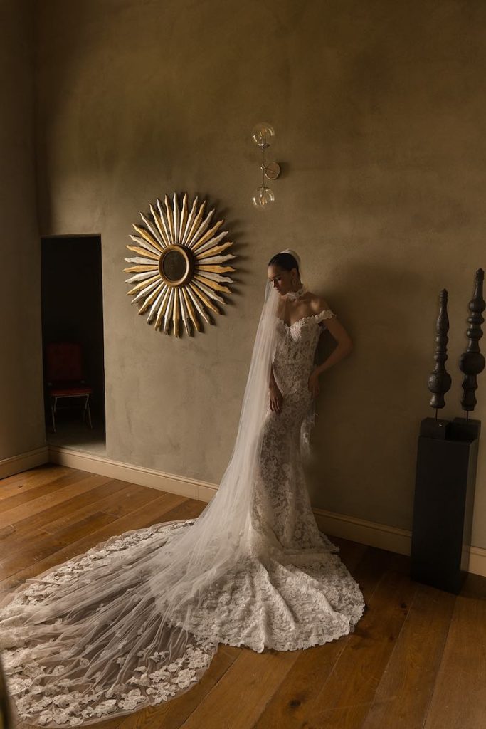 In questa foto, una modella posa indossando un abito da sposa a sirena in pizzo Donatella Gallo 2027 con scollo omerale in pizzo con lungo velo abbinato