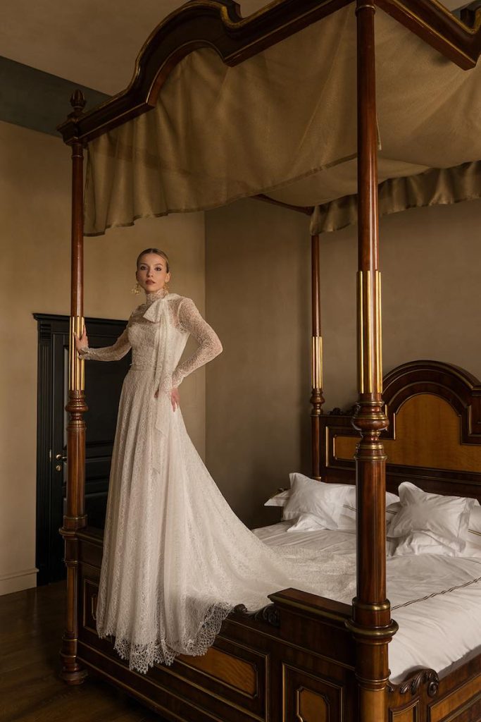 In questa foto, una modella posa in piedi sul bordo di un letto indossando un abito da sposa scivolato Donatella Gallo 2027 in pizzo con maniche lunghe e collo alto fermato da un grande fiocco