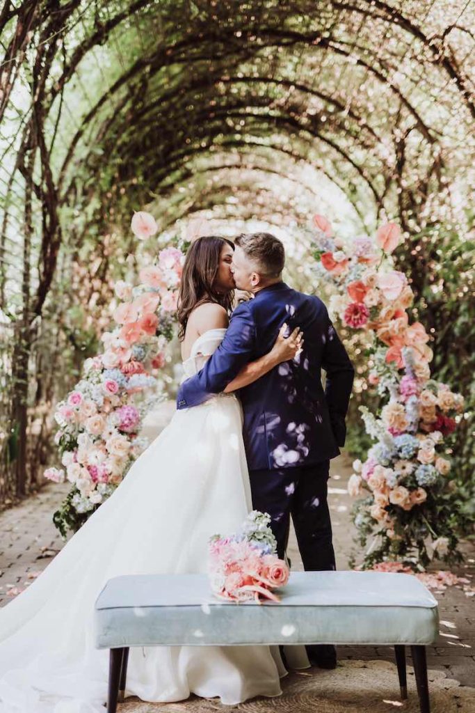 In questa foto, due sposi si baciano al termine del loro matrimonio davanti ad un arco floreale multicolor