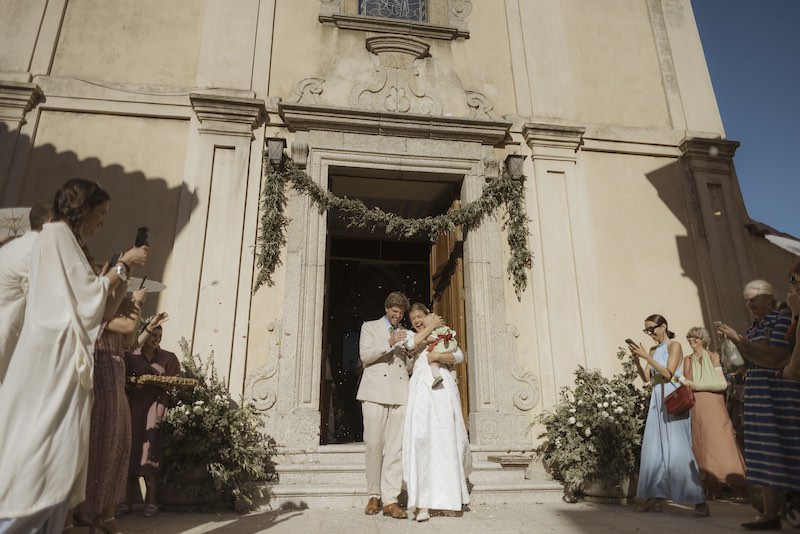 In questa foto, gli sposi Camilla e Niccolò con in braccio la loro bimba all'uscita della chiesa durante il lancio del riso. Dietro di loro un festone di fiori e ulivi e composizioni laterali
