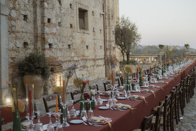 In questa foto, un lungo tavolo imperiale allestito da Elena Bitonte con tovagliato color ruggine, vasetti di colore verde, fiori multicolor, candele, tovaglioli e tamburelli en pendant