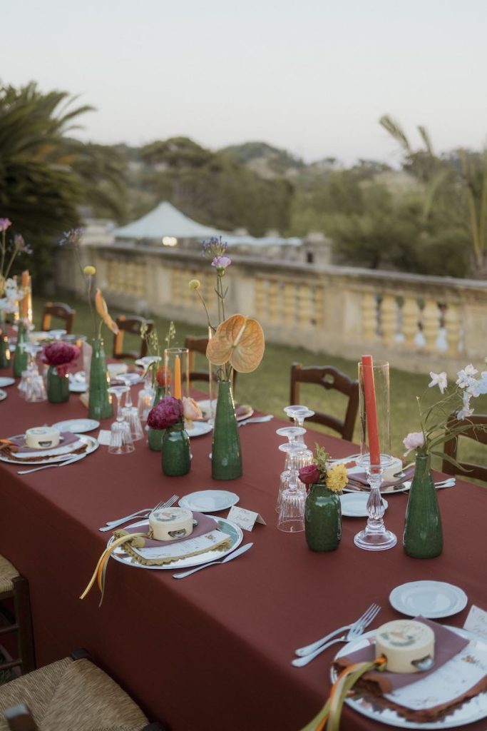 In questa foto, il dettaglio di un lungo tavolo imperiale allestito da Elena Bitonte con tovagliato color ruggine, vasetti di colore verde, fiori multicolor, candele, tovaglioli e tamburelli en pendant