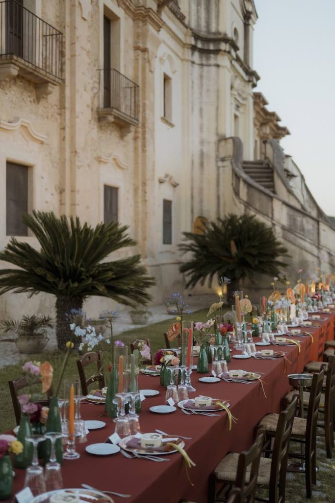 In questa foto, un lungo tavolo imperiale allestito da Elena Bitonte con tovagliato color ruggine, vasetti di colore verde, fiori multicolor, candele, tovaglioli e tamburelli en pendant