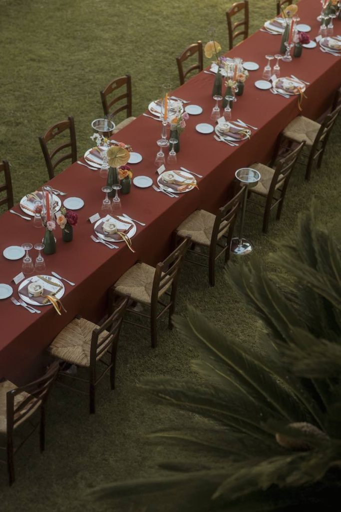 In questa foto dall'alto, un lungo tavolo imperiale allestito da Elena Bitonte con tovagliato color ruggine, vasetti di colore verde, fiori multicolor, candele, tovaglioli e tamburelli en pendant