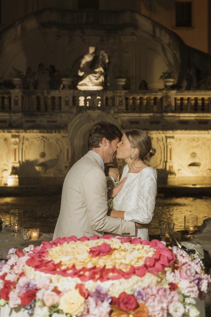 In questa foto, due sposi si baciano dietro alla loro torta nuziale ad un piano decorata con petali e fiori multicolor