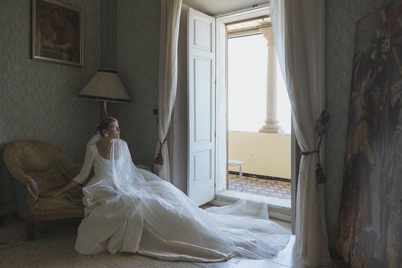 In questa foto, una sposa sdraiata su un divano mostra il suo abito con lungo velo, entrambi disegnati da lei e cuciti e ricamati nel suo atelier Le Tre Sarte