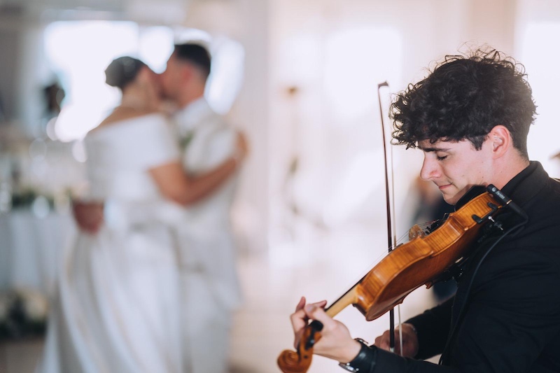 In questa foto, due sposi sullo sfondo ballano un lento sulle note di un violinista - in primo piano - del team di Direzioni Parallele