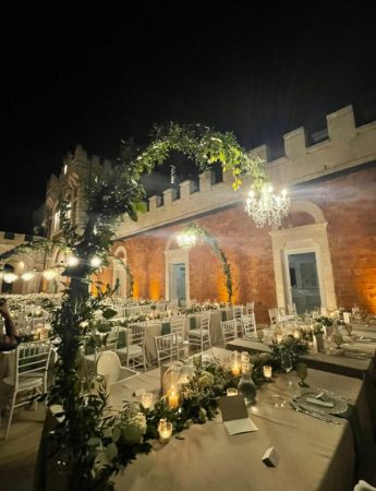 In questa foto l'allestimento di un ricevimento di nozze all'aperto sulla terrazza di un castello nei toni del bianco e del verde salvia. Ogni tavolo rettangolare sistemato uno accanto all'altro è illuminato da lanterne a braccio ricoperte da foglie che riprendono i runner. Le tovaglie sono rosa cipria mentre i tovaglioli pendenti verde salvia