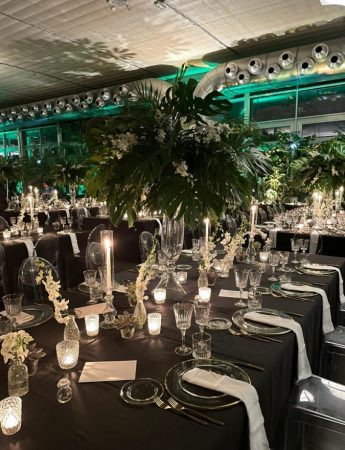 In questa foto l'allestimento di un matrimonio in un capannone industriale nei toni del nero, dell'argento e del verde. Sul tetto si distinguono i condotti dell'aria in alluminio. La mise en place è in cristallo. Al centro dei tavoli imperiali sono presenti dei vasi con palme e fiori bianchi