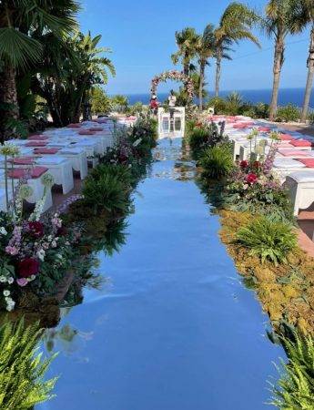 In questa foto l'allestimento di un matrimonio civile sul mare alla luce del mattino. Il corridoio che conduce all'arco delle nozze è realizzato con un materiale riflettente. Lungo tutto il corridoio sono presenti foglie e fiori bordeaux, viola e rosa. I posti degli ospiti sono realizzati con delle panche bianche su cui sono poggiati dei cuscini bordeaux e rosa. Tutta la location è circondata da palme