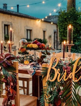 In questa foto il tavolo degli sposi decorati in stile Boho Chic con palme e fiori rossi e gialli. Dietro le sedie degli sposi sono appese le scritte"Bride" e "Groom"