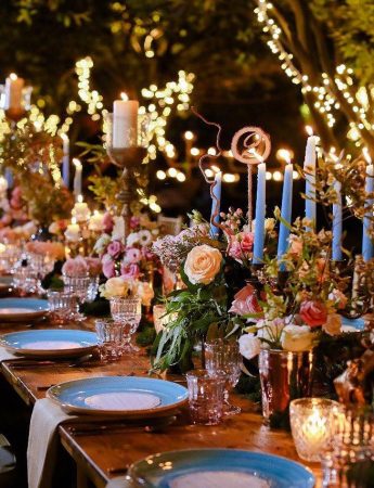 In questa foto una mise en place in stile Boho Chic su un tavolo imperiale in legno. Il runner è realizzato con candele azzurre, fiori colorati e vasetti luminosi. L'allestimento è completato con piatti azzurro polvere