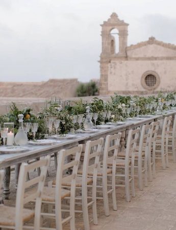 In questa foto un allestimento Shabby Chic nel cortile di una chiesa di campagna. Le sedie sono bianche e delle composizioni di foglie e fiori sono usate come runner.