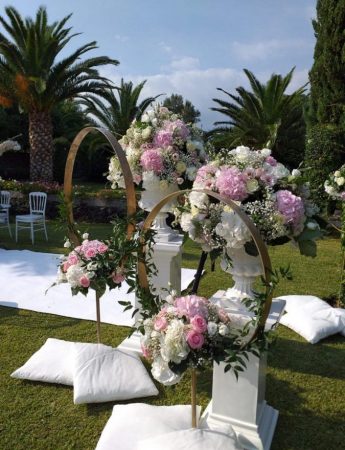 In questa foto l'allestimento floreale per una cerimonia civile su un prato. Felce e fiori rosa e bianchi decorano dei cerchi dorati sospesi e dei vasi bianchi. Sul prato sono disposti dei cuscini bianchi e un tappeto coordinato