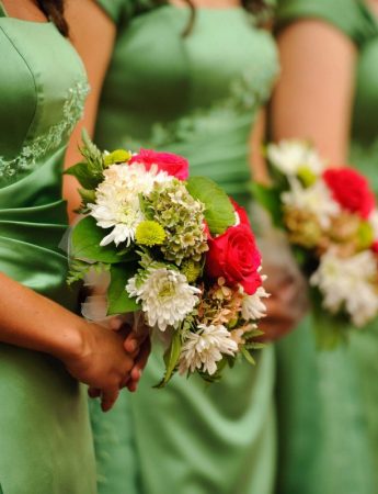 In questa foto 3 damigelle d'onore con un abito verde brillante tengono tra le mani bouquet di girasoli bianchi e rossi