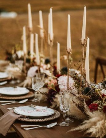 In questa foto una mise en place in stile Boho Chic per un ricevimento di nozze su un campo di grano. Un tavolo imperiale di legno visto in obliquo è decorato con candele alte e bianche, fiori rossi e sottopiatti in paglia